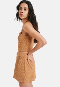 Femme aux longs cheveux bruns, portant un haut en crochet sans manches de couleur beige assorti et une jupe taille haute, posant avec une main sur la hanche.