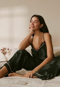 Musta kukallinen pyjama-asusetti, jossa on pitsireunus ja vaaleanpunaiset yksityiskohdat, sisältäen hihattoman topin ja väljemmät housut, esillä sängyllä.