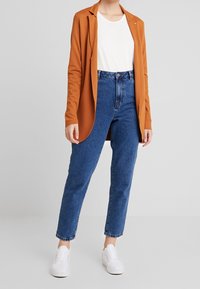 Cardigan lungo arancione, t-shirt bianca, jeans blu a vita alta e sneakers bianche. Il cardigan ha un davanti aperto e non presenta chiusure.