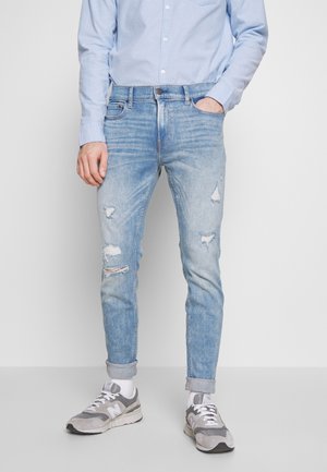 Homme portant un jean bleu clair déchiré, une chemise bleu clair boutonnée, des chaussettes blanches et des baskets grises, une main dans la poche.