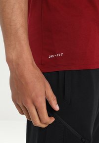 Camiseta Dri-FIT color burdeos con una textura suave. Presenta un logo blanco cerca del dobladillo y se combina con pantalones deportivos negros que tienen un bolsillo lateral.