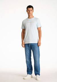 Ljusblå kortärmad t-shirt med texten "Köpenhamn" i en ljusare nyans, kombinerad med blå jeans och vita sneakers. Enkelt och avslappnat design.