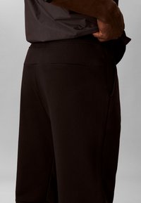 Άνδρας που φοράει μαύρο παντελόνι με ελαστική μέση και πλαϊνές τσέπες, ρυθμίζοντας ένα γκρι πουκάμισο περασμένο μέσα από το παντελόνι μπροστά σε ένα μονόχρωμο φόντο.