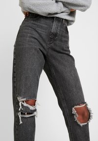 Černé džíny s vysokým pasem, které mají roztržení na kolenou a potrhané zakončení. Obsahují klasický 5-kapsový design a slabou texturu.