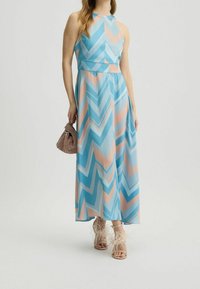 Robe maxi à motif zigzag bleu et rose, sans manches et avec un col haut. Portée avec des sandales à talons ornées de plumes et un sac à main texturé.