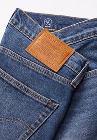 Blue denim jeans with a brown leather back patch. Features orange stitching and a five-pocket design. Material is soft with a slightly worn texture.