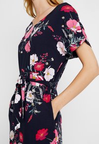 Blumenkleid in Marineblau mit lebhaften pinken und weißen Blumen, kurzen Ärmeln, einer taillierten Taille mit Bindegürtel und seitlichen Taschen.