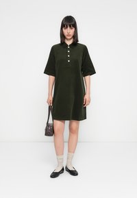 Olivengrønn, kortermet polo kjole med knapper, glatt tekstur, knelange, kombinert med beige sokker og svarte leiligheter, holder en liten brun veske.