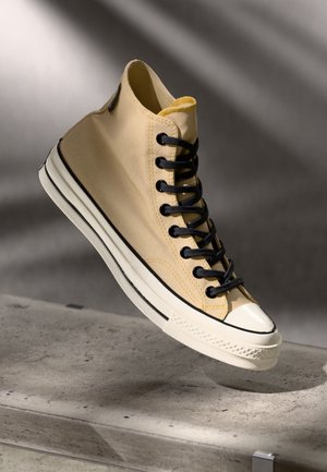 Zapatilla alta de lona beige claro con cordones negros, suela de goma blanca y detalles negros. Acentos amarillos en la zona del collar.
