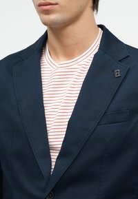 Άνδρας που φοράει μπλε σκούρο σακάκι με μια μικρή ορθογώνια μαύρο-λευκή καρφίτσα στο πέτο και ένα λευκό πουκάμισο με λεπτές κόκκινες ρίγες.