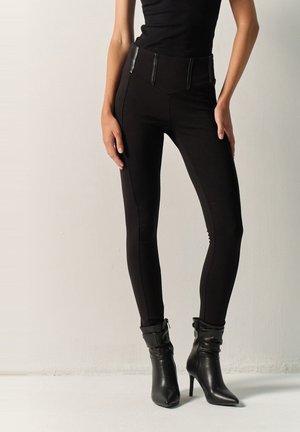 Mujer con pantalones ajustados de cintura alta en negro con paneles de piel sintética, combinados con botines negros de tacón y una camiseta sin mangas negra.