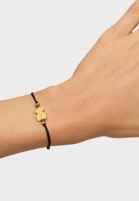 Pulsera de cordón negro con un charm de oso dorado de bordes suaves, sobre un fondo en tono de piel neutro. Diseño simple con longitud ajustable.