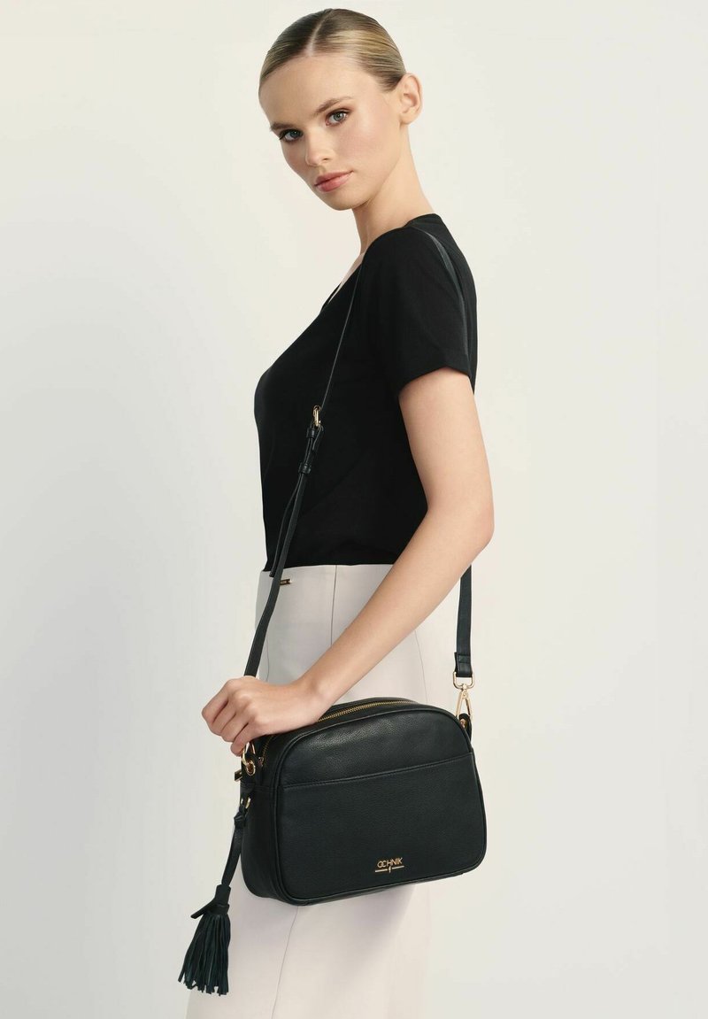 Czarna skórzana torba na ramię o wyraźnym kształcie, z złotymi detalami i frędzlem na suwaku. Miękka, teksturowana powierzchnia.