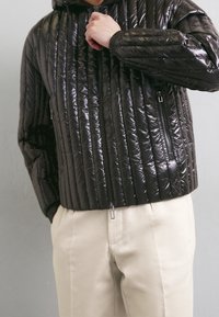 Vadderad svart jacka med en glänsande yta, med vertikala kviltade linjer, en framzipper och sidofickor. Bärs över krämfärgade byxor.