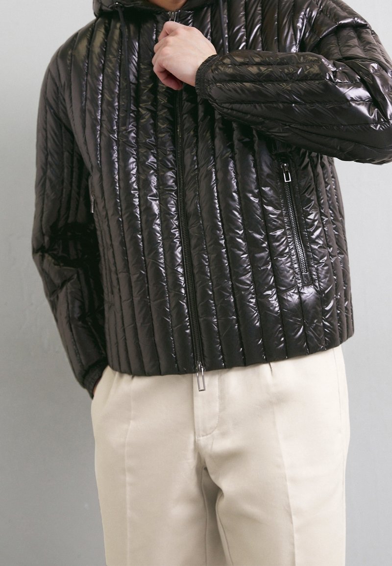Vadderad svart jacka med en glänsande yta, med vertikala kviltade linjer, en framzipper och sidofickor. Bärs över krämfärgade byxor.