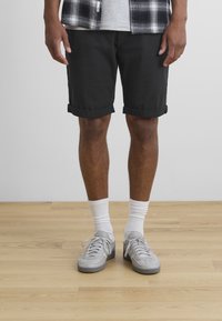 Svarte shorts med rullet kant, parret med lysegrå sneakers og hvite ankelsokker, stående på et trebord mot en ensfarget bakgrunn.