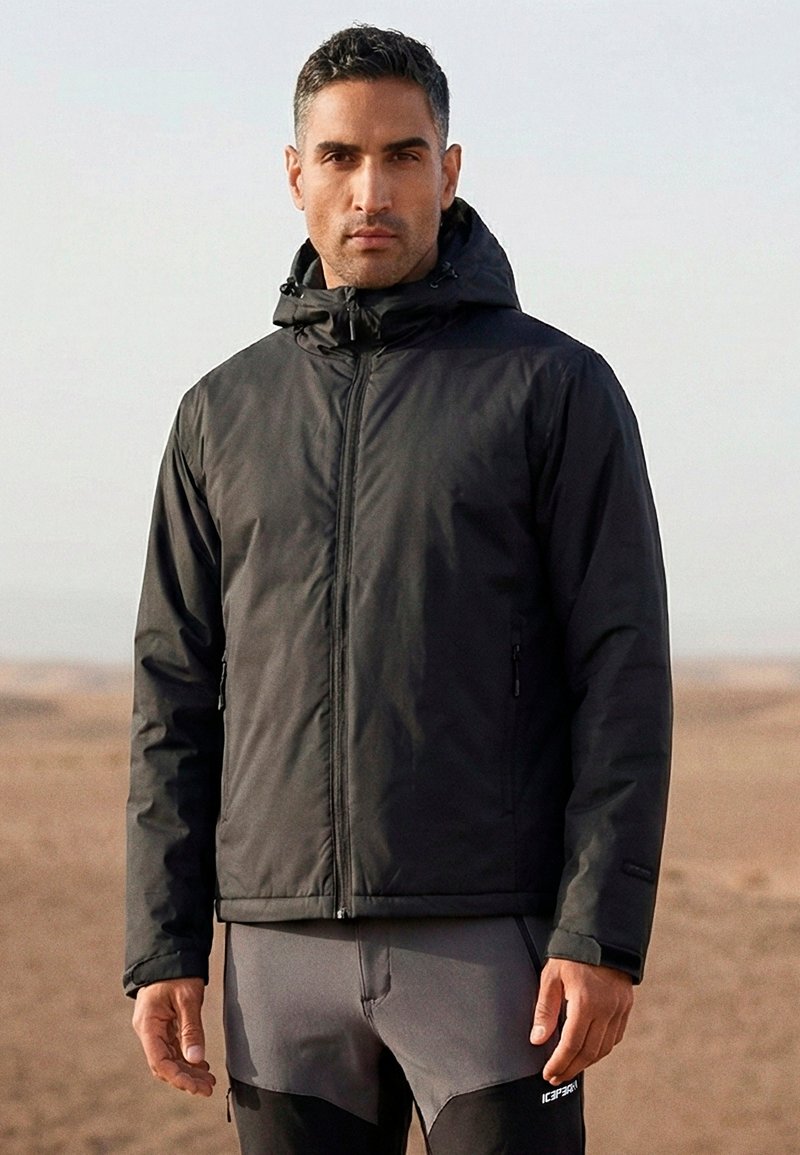 Homme portant une veste noire à capuche et un pantalon de sport gris, debout à l'extérieur dans un paysage aride.