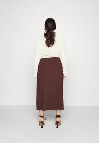 Bruine gebreide rok met een aansluitende taille en een rechte silhouet. Zwarte hakken met enkelbandjes. Model wordt van achteren getoond.