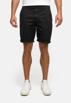 Mann trägt schwarze Shorts mit umgeschlagenem Saum und weiße Sneakers, steht mit entspannten Armen vor einem schlichten hellen Hintergrund.