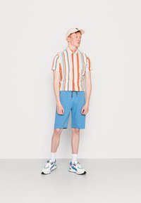 Stripet skjorte med kort erm i oransje, grønn og hvit; blå shorts; hvite joggesko med fargerike aksenter; beige caps.