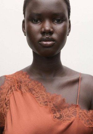 Joven mujer negra con cabello corto, vistiendo una blusa de satén color naranja óxido con encaje, sobre un fondo blanco liso.