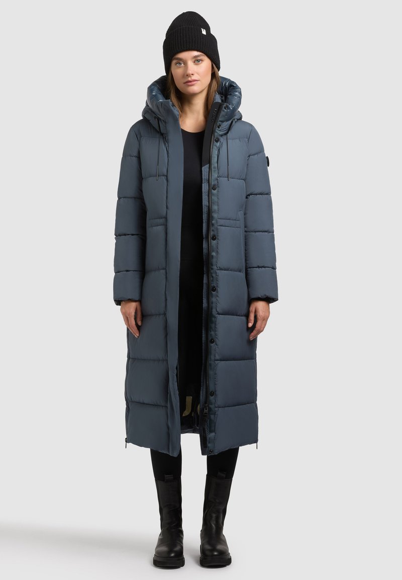 Tamsiai mėlyna, paminkštinta ilga striukė su gobtuvu, priekiniais mygtukais ir šoninėmis užtrauktukais. Užsivilkta ant juodų drabužių ir juodų batų.