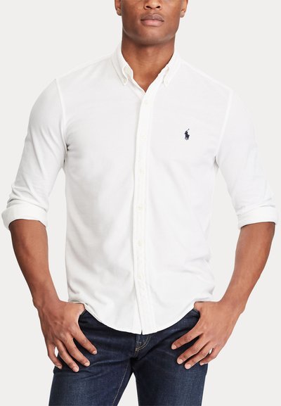 Homme portant une chemise blanche à boutons avec manches retroussées et un jean foncé, les mains posées sur les hanches, sur un fond uni.