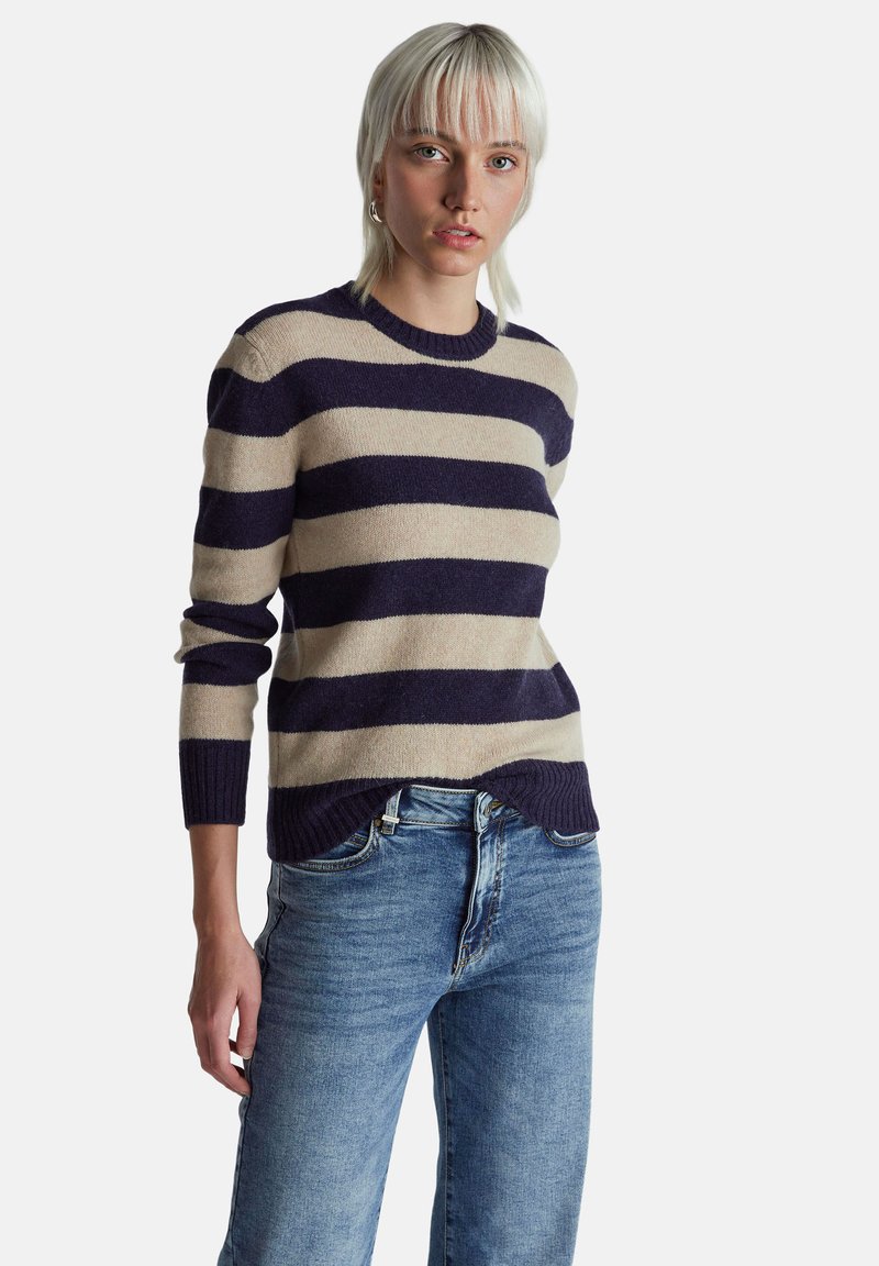 Kobieta o jasnoblond włosach, ubrana w granatowo-beżowy sweter w paski i niebieskie dżinsy, stojąca na tle jednolitego białego tła.