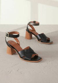 Schwarze Leder-High-Heels mit Kreuzriemen, quadratischer offener Zehenpartie, strukturiertem braunem Blockabsatz und verstellbarem Knöchelriemen mit goldener Schnalle.