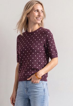 Lyshåret kvinde smilende og kigger mod højre iført en lilla kortærmet sweater med hvide polkaprikker og lyseblå jeans, hånd i lommen.