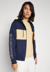 Námornícky modrá mikina na zips s béžovými prsiami a veľkým logom na rukáve. Obsahuje kapucňu so šnúrkou a predné vrecká.