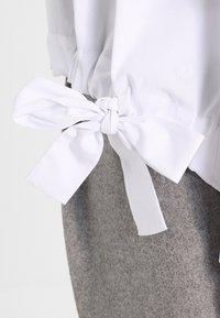 Blouse blanche avec un nœud noué sur le côté. Tissu doux, détail froncé à l'ourlet, associée à un vêtement texturé gris clair en dessous.