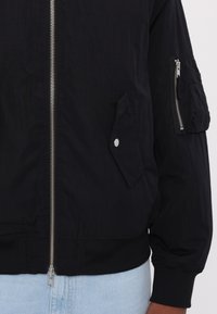 Black bomber jacket with a front zipper, ribbed hem, side pockets, and a zippered arm pocket; made from smooth, lightweight fabric.