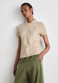 Beige polo skjorte med krage og tre knapphull, laget av bomull, med en liten blå logo på venstre bryst. Kombinert med grønne bukser.