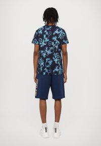 Navy t-shirt z modro in turkizno cvetličnim vzorcem, v kombinaciji z navy kratkimi hlačami z logotipom "VERSACE JEANS" v zlati barvi na strani.