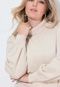 Maglione beige a maglia con colletto alto e maniche a sbuffo. Texture liscia con polsini a coste. Orecchini a cerchio dorati, molto evidenti, arricchiscono il look.