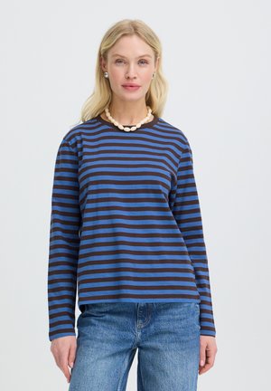 Femme blonde portant une chemise à manches longues rayée bleu et noir, un jean bleu, un collier de perles et des boucles d’oreilles argentées sur un fond uni.