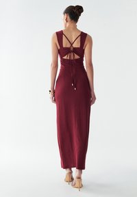 Bordo spalvos maxi suknelė iš ribbingo audinio turi iškirptę nugaroje su kertamomis petnešėlėmis ir raištelių detalę, lygią tekstūrą ir aptemptą siluetą.