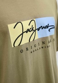 Oliven grøn T-shirt med et rektangulært grafisk motiv i gul og lys blå, med "JackJones" i sort kursiv og teksten "ORIGINALS WORLDWIDE" nedenunder.