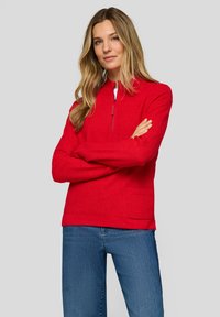 Frau mit langen blonden Haaren, die einen roten Pullover mit Viertelreißverschluss und Tasche, blaue Jeans trägt und mit verschränkten Armen vor einem grauen Hintergrund steht.