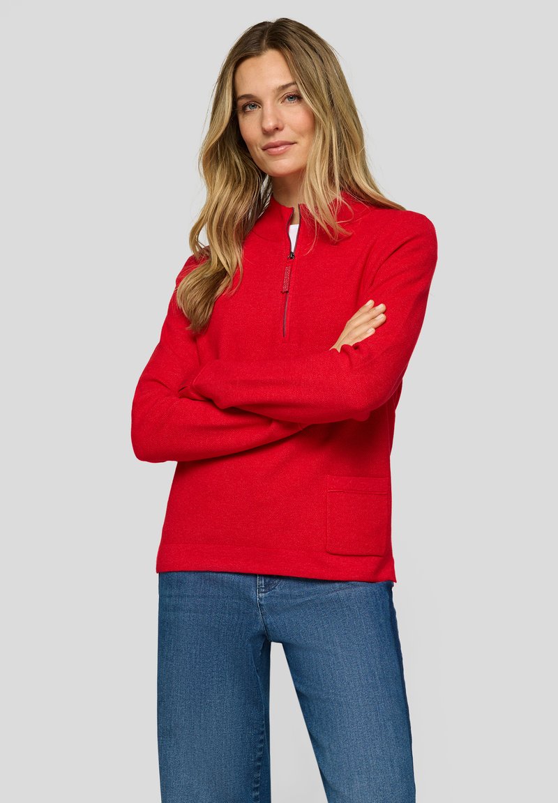 Frau mit langen blonden Haaren, die einen roten Pullover mit Viertelreißverschluss und Tasche, blaue Jeans trägt und mit verschränkten Armen vor einem grauen Hintergrund steht.