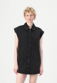 Zwarte denim jurk met korte mouwen, knoopsluiting aan de voorkant en een borstzak. De stof heeft een gladde textuur en een klassiek kraagontwerp.
