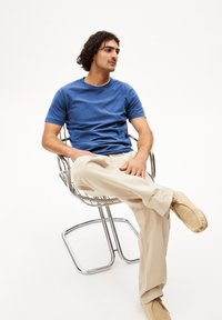 Blauwe katoenen T-shirt met een ronde hals, gecombineerd met beige broeken. Het model zit met gekruiste benen in een chromen draadstoel en draagt lichte schoenen.