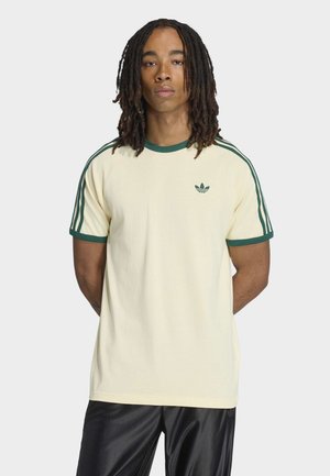 Muž s dredy po ramena, který nosí krémové tričko Adidas se zelenými pruhy a logem, v kombinaci s lesklými černými kalhotami, na bílém pozadí.