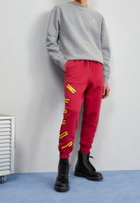 Felpa grigia con maniche lunghe e un piccolo logo. Pantaloni della tuta rossi con scritte gialle e anfibi neri con lacci, texture minima.