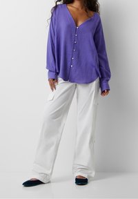 Mujer vistiendo una blusa morada de botones suelta, pantalones cargo blancos de pierna ancha y zapatillas oscuras, de pie contra un fondo blanco sencillo.