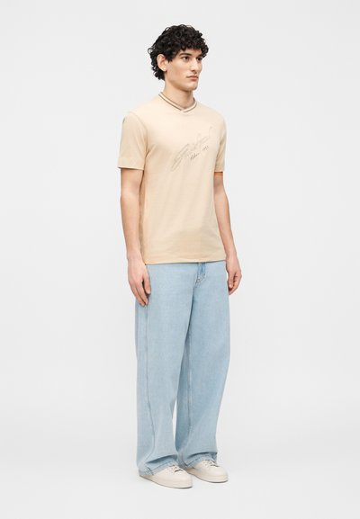 Νεαρός με σγουρά μαλλιά που φοράει μπεζ κοντομάνικο πουκάμισο, ανοιχτό μπλε τζιν με φαρδιά μπατζάκια και λευκά αθλητικά παπούτσια, ο οποίος στέκεται μπροστά σε απλό φόντο.