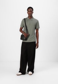 Menns kortermet oliven grønn polo med glidelås krage, kombinert med vide svarte bukser. Holder en liten svart crossbody-veske. Hvite sneakers.