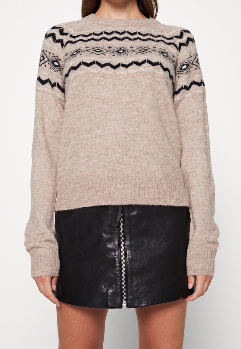 Femme portant un pull en maille beige avec des motifs géométriques noirs et une mini-jupe en cuir noire avec une fermeture éclair à l'avant.