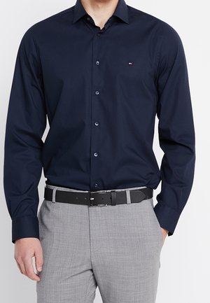 Homme portant une chemise bleu marine à boutons rentrée dans un pantalon de costume gris clair avec une ceinture noire, une main dans la poche.